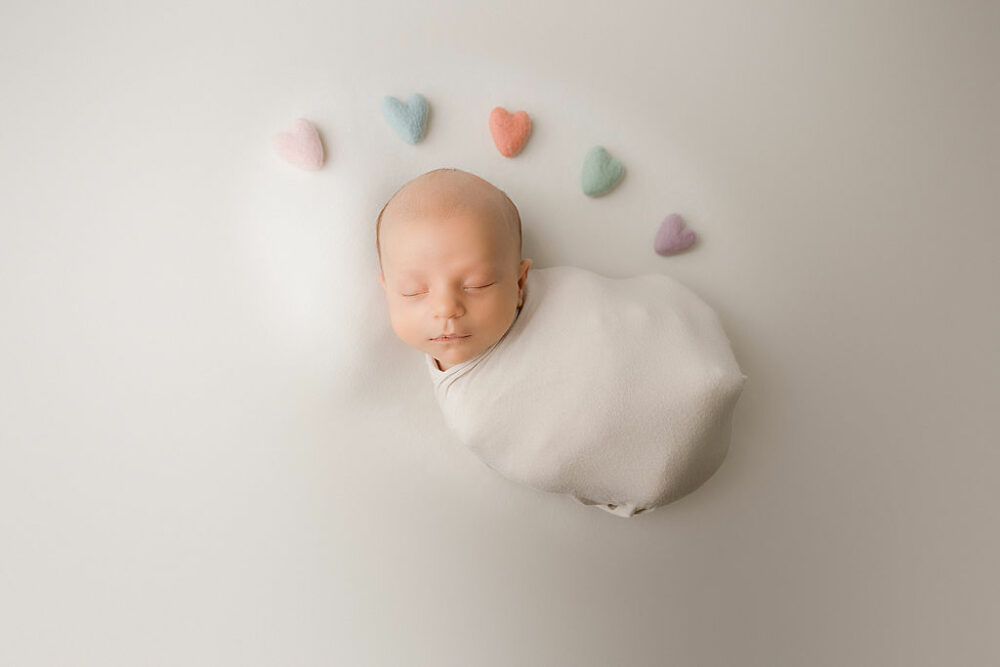 A newborn sleeping on back, wrapped in swaddle and adorned with multicolored, felt hearts posing for his baby photo studio newborn session in Westampton, New Jersey.