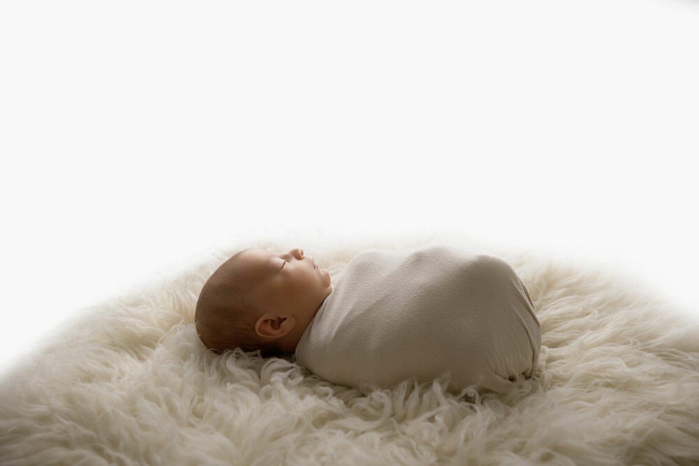 Side view of a sleeping baby that is wrapped and laying on bean bag photography prop on top of textured blanket for an in-studio navy blue newborn session in Southampton, New Jersey.