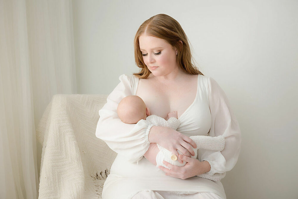 Woman sitting and holding her newborn as she looks down at her, breastfeeding in a professional studio for her white and green newborn session in southampton, New Jersey.