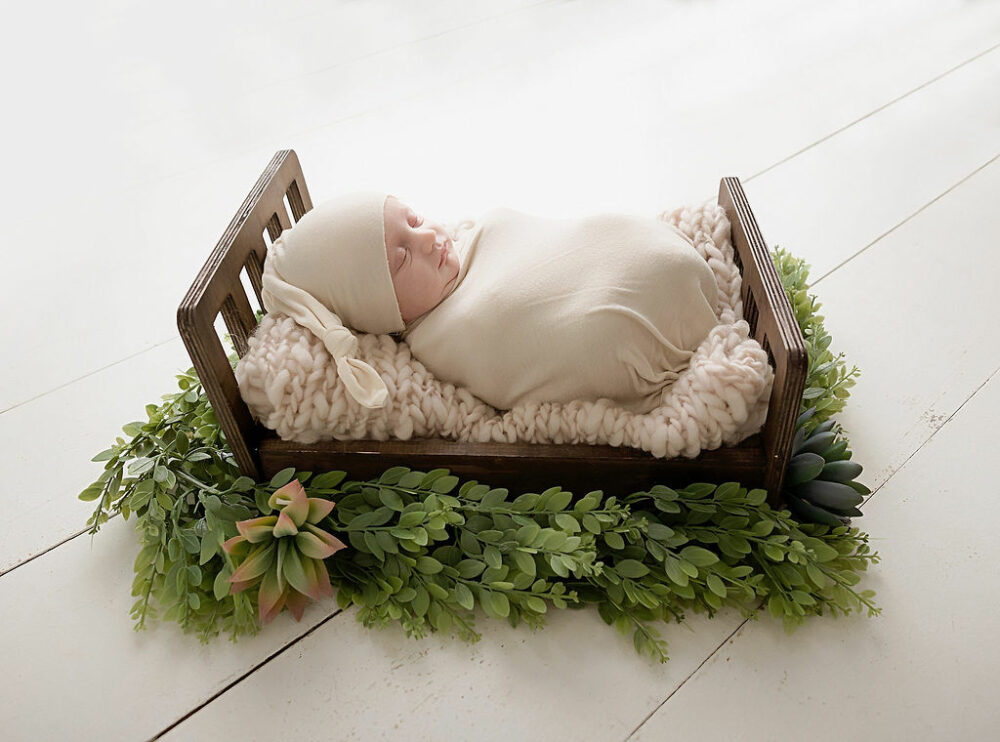 A sleepy infant sleeping on textured blanket and tiny photography pop crib adorned with greenery for a white and green newborn session in profession photography studio in Hamilton, New Jersey.