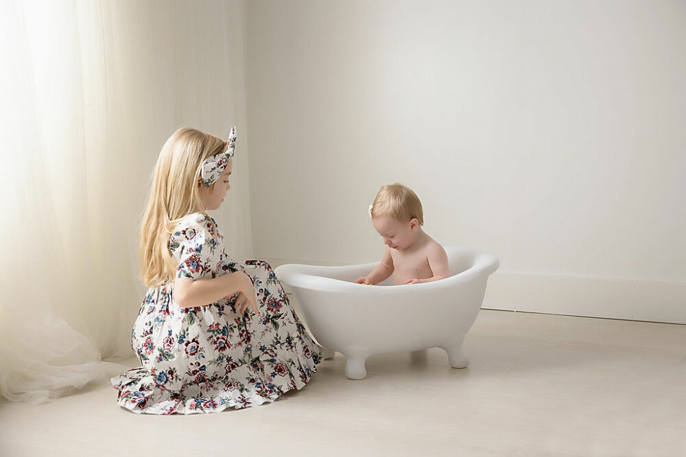 A toddler sitting in bathtub, photography prop, and playing in the water as her sister beside her for their first birthday ideas taken in professional studio for her baby milestones session in Southampton, New Jersey.