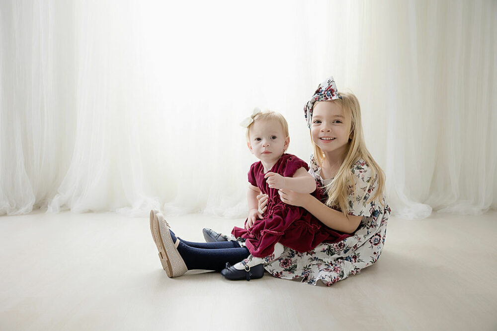 A big sister sitting on ground, carrying her toddler sibling and smiling for their sibling portraits taken during her winter wonderland first birthday session in Hamilton, New Jersey.