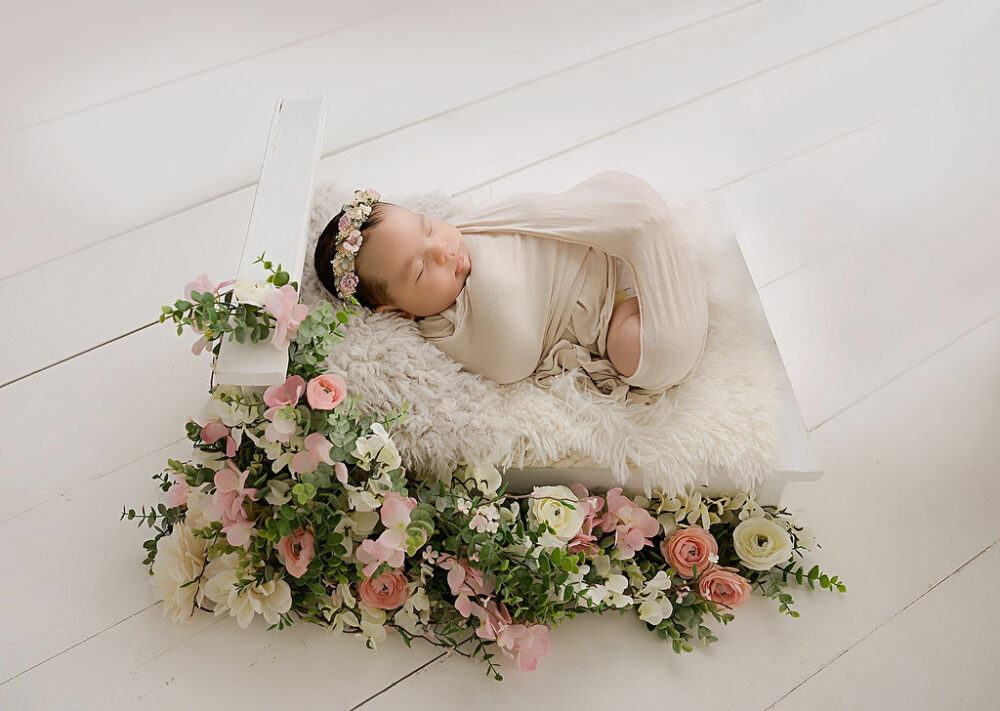 Newborn portrait of a girl wrapped in swaddle sleeping on textured blanket on tiny crib adorned with flowers for her three month newborn photography session in Southampton, New Jersey.
