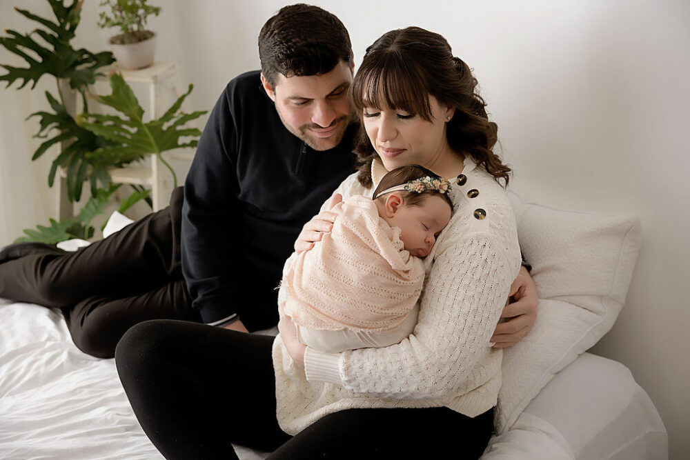 A man and woman looking at their infant daughter as they sit on bed photography prop wearing sweaters and pants for their newborn infant photography session in Medford, New Jersey.