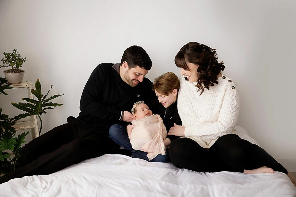 Family portrait of a couple and their two children sitting in bed photography set wearing sweaters as they look lovingly at their infant daughter for her three month old newborn session in Deptford, New Jersey.