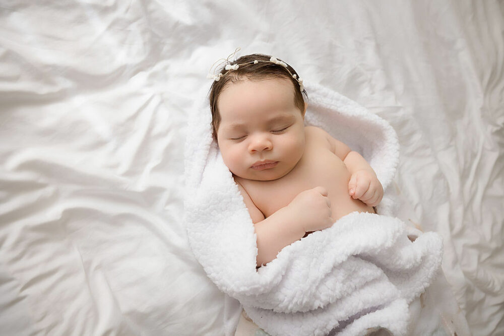 An infant girl sleeping on her back and cozy in a blanket wearing a cute headband during her three month old newborn session in Camden, New Jersey.