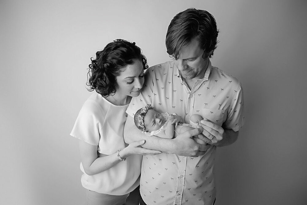 Black and white photo of a man and a woman posing and looking down at their infant daughter as her father holds her in his arms during their themed newborn photography session in Westampton, New Jersey.