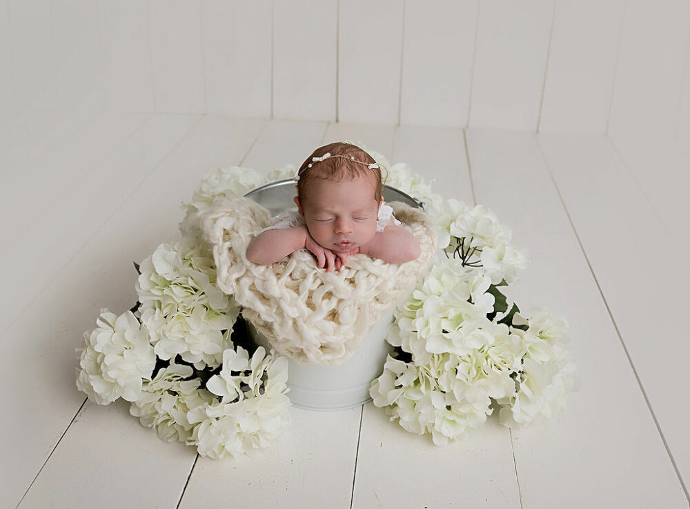 A infant portrait of a girl sleeping in a bucket adorned with white flowers and a textured blanket posed for her themed newborn session in Hamilton, New Jersey.
