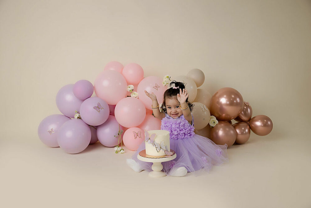 A toddler girl sitting on floor in front of a cake with large balloon garden in the backdrop adorned with flowers and butterflies for her butterfly first birthday session in Westampton, New Jersey.