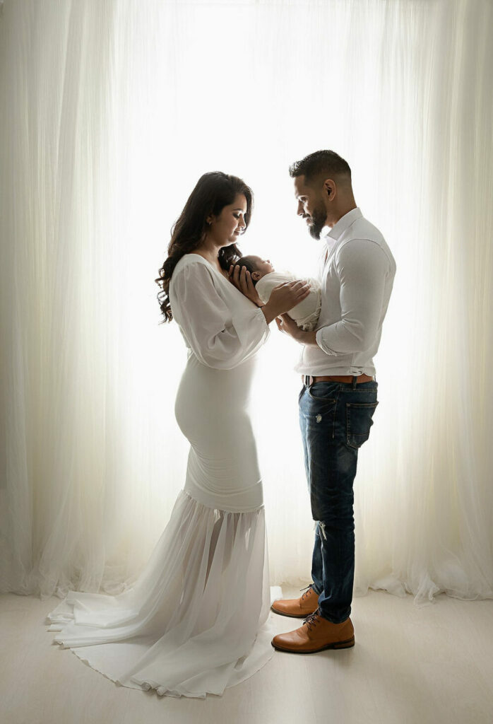 A couple maternity photos of a man and woman facing each other and holding their son in between, them standing against a light and bright background for an Indian inspired newborn session in West Hampton, New Jersey.