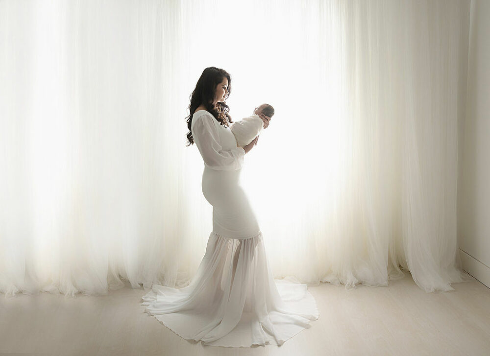 Family portrait of a woman standing to the side as she holds her newborn son posing for her newborn photo shoot session in Cherry Hill, New Jersey.