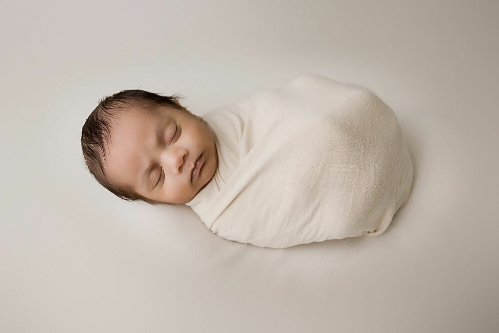 An infant sleeping on his back, wrapped in swaddle against a light backdrop for his professional baby pictures taken during his Indian newborn session in Southampton, New Jersey.