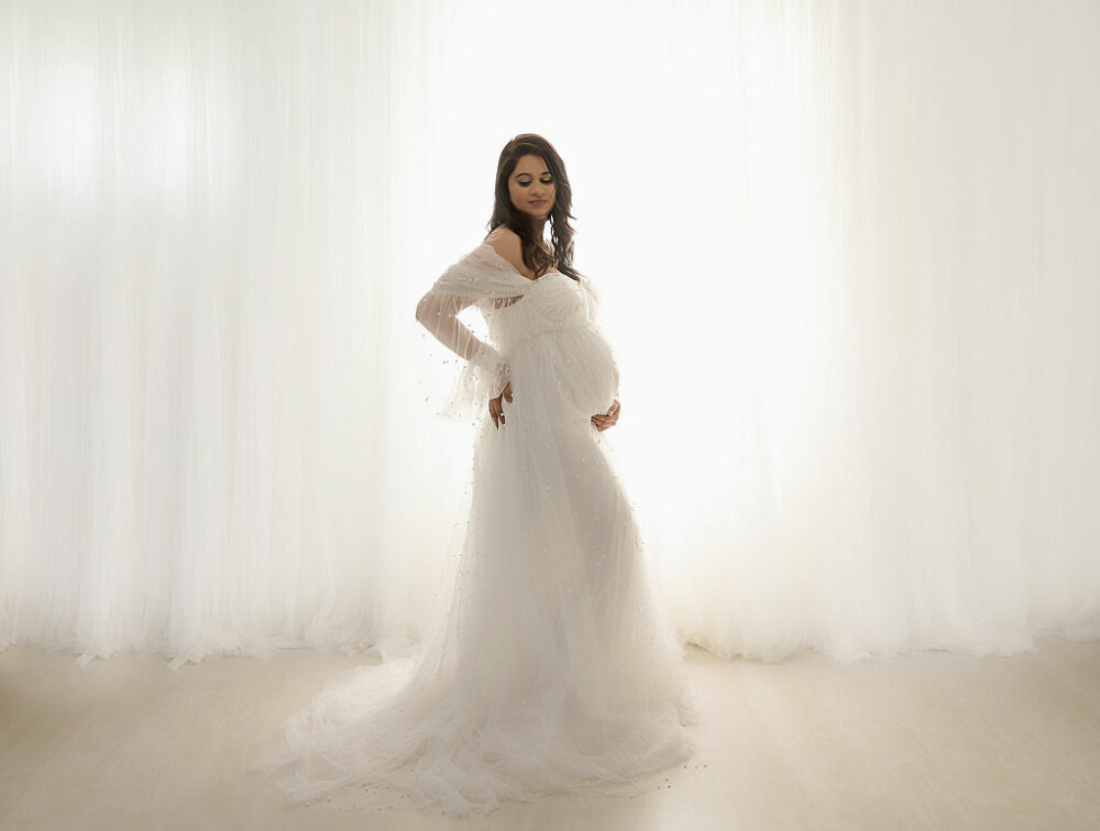 A pregnant woman standing against a light and bright background oh wearing long maternity gown and holding her belly for her maternity session in Camden, New Jersey.