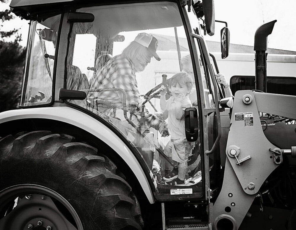 Black-and-white photo of a man sitting in a tractor with his grandson in side, taken during a farm lifestyle family session in Deptford, New Jersey.