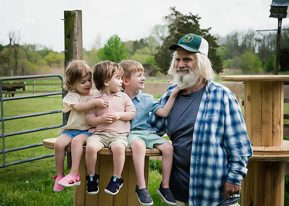A man standing and posing with his three grandchildren as they sit and touch his gray beard during a family session at their family farm in Westampton, New Jersey.