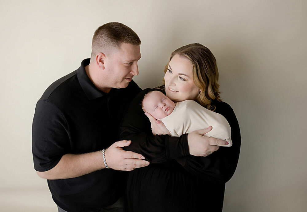 A man and a woman looking at each other as she holds her infant son in her arms wearing matching colored clothing for their newborn photography session in Eastampton, New Jersey.