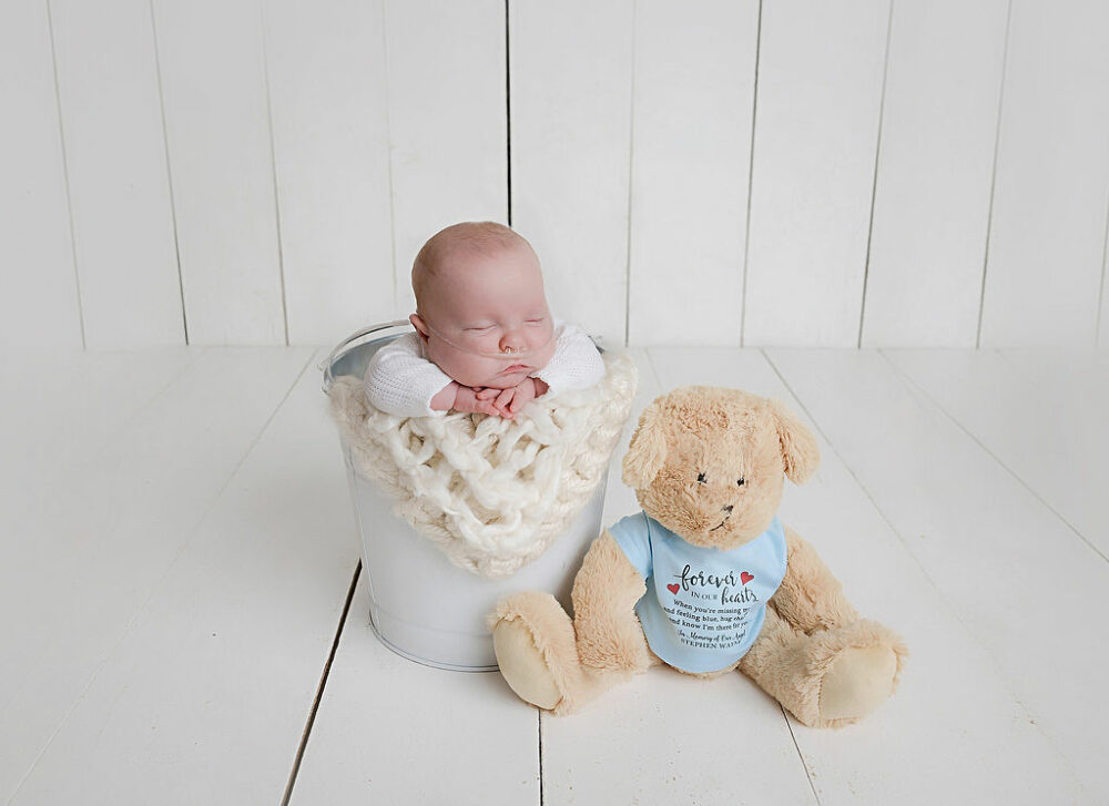 Newborn portrait of a boy posed in a bucket lined with textured blanket and a teddy bear plushy sitting next to him for his twin Nicu newborn session in Cherry Hill, New Jersey.