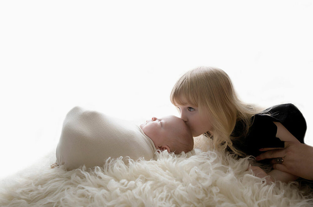 A sibling portrait of a toddler sister kissing her infant brother as he sleeps on a textured blanket for his twin Nicu newborn session in Mount Holly, New Jersey.