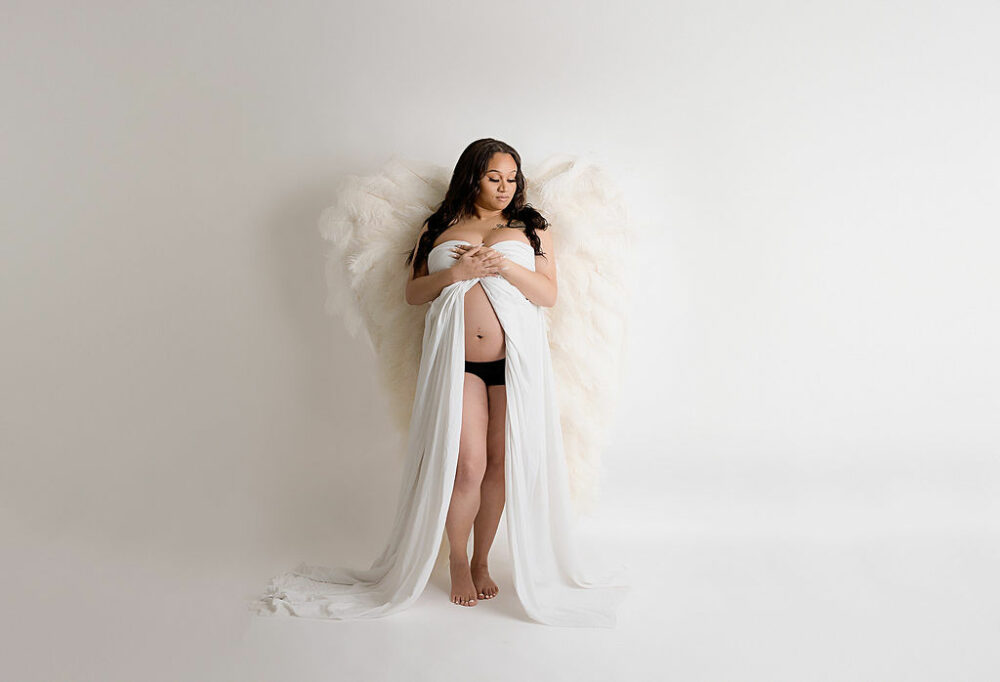 A woman standing and posing with her belly showing as she wears white gown with large angel wings for her angelic in studio maternity photography session in Southampton, New Jersey.