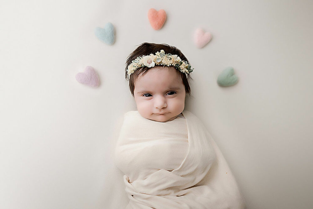 An infant portrait of a girl resting on her back as she is swaddled, wearing headband and alert with hearts surrounding her head for her floral Newborn session in Southampton, New Jersey.