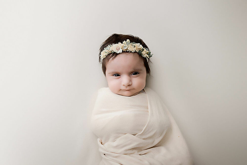 A infant portrait of a girl awake and resting on her back, wearing headband and swaddled for her in-studio floral newborn session in Cherry Hill, New Jersey.