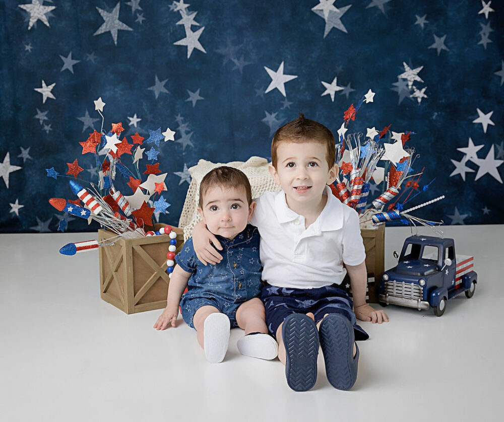 Two boys sitting and hugging each other, and smiling against starry backdrop in professional studio for USA holiday mini sessions is Camden, New Jersey.