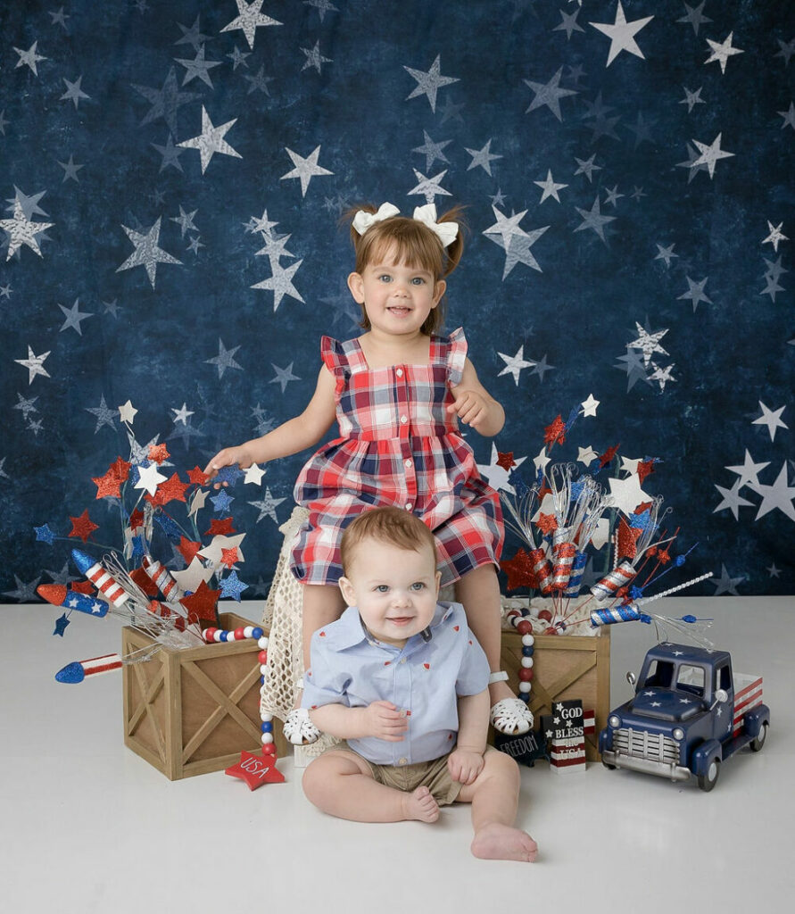 A toddler girl sitting with her younger brother, smiling and having a good time during her USA holiday mini sessions in Medford, New Jersey.