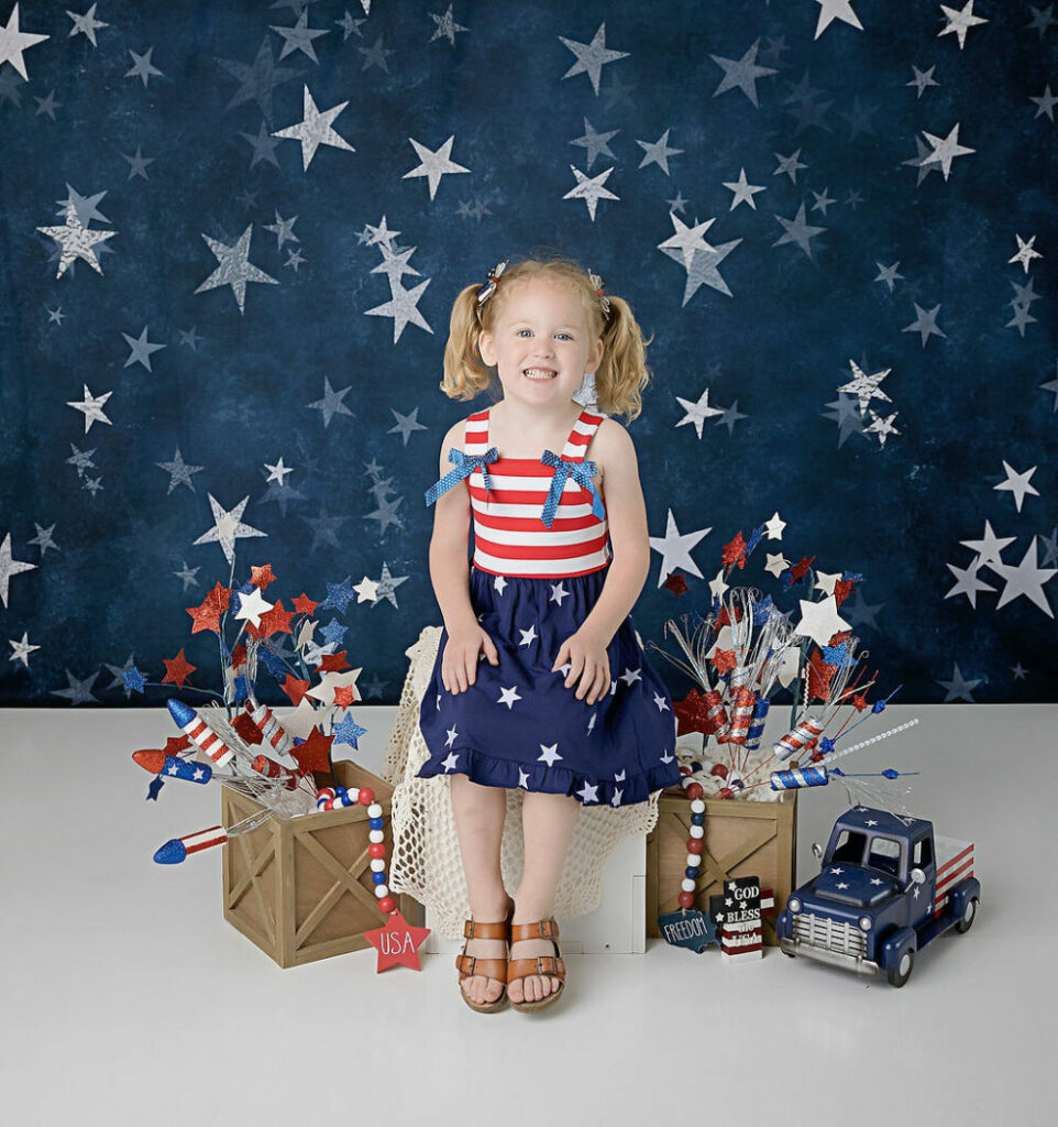 A themed photograph of a girl, smiling, wearing cute dress and sitting in a Fourth of July themed set for professional, USA holiday minis in Deptford, New Jersey.
