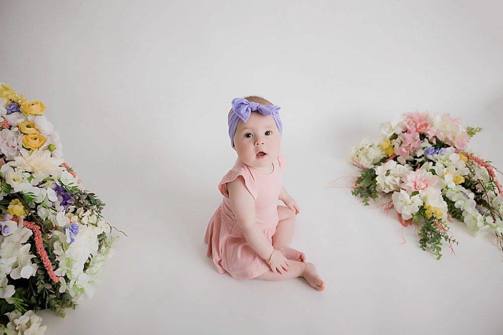 A infant portrait of a girl wearing cute overalls and headband, sitting next to an assortment of flowers as props for her professional photographer portraits taken in Tabernacle, New Jersey.