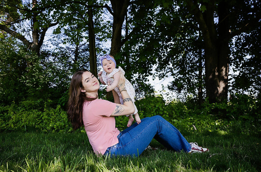 A woman sitting outdoors in the grass in front of lush greenery holding her daughter up in the air for photography ideas in Eastampton, New Jersey.