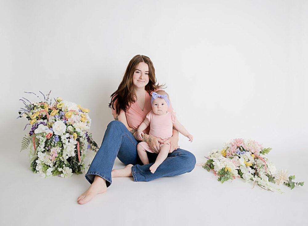 A woman and her infant daughter sitting on the floor next to beautiful flower assortment wearing like colors in causal clothing for a In-Studio Mother’s Day session in Southampton, New Jersey.