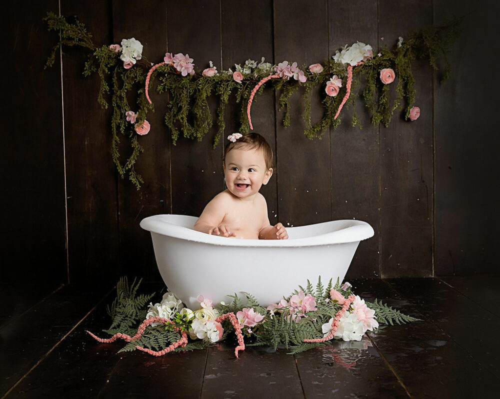 A birthday portrait of a toddler girl smiling in a bathtub photography prop with a brown backdrop and greenery taken during her mermaid first birthday session in Tabernacle, New Jersey.