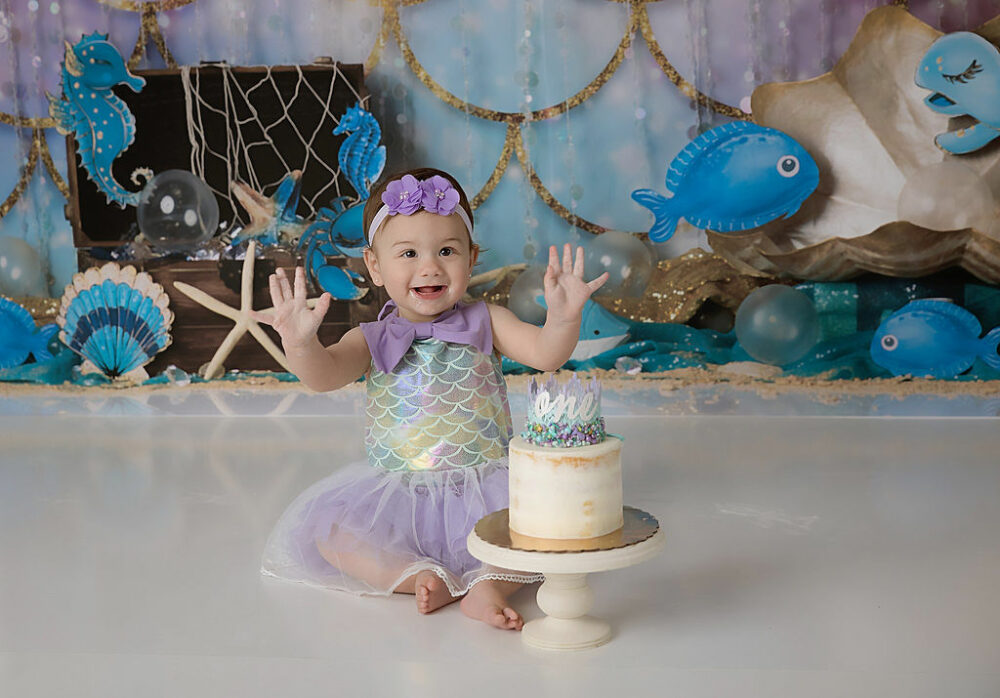 A close-up of a toddler girl smiling and wearing cute dress next to a cake for her mermaid themed first birthday party ideas in Deptford, New Jersey.