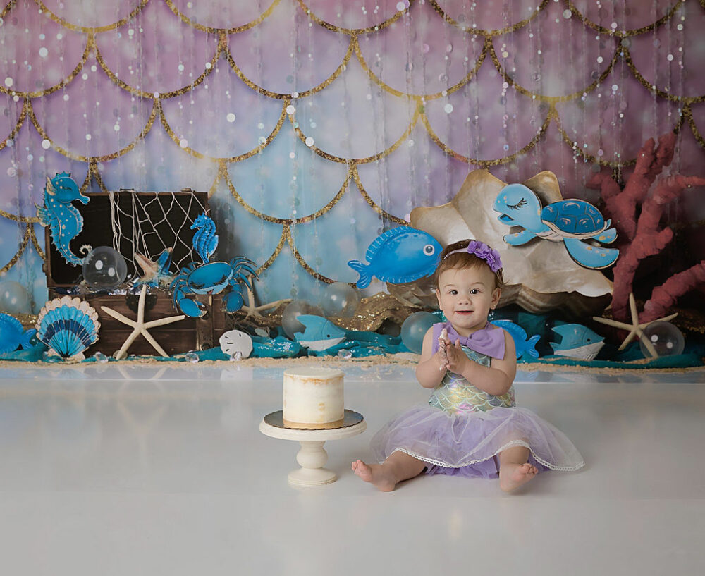 A cute picture of a toddler girl sitting in front of mermaid theme backdrop with a plain cake smiling for her mermaid first birthday session in Medford, New Jersey.