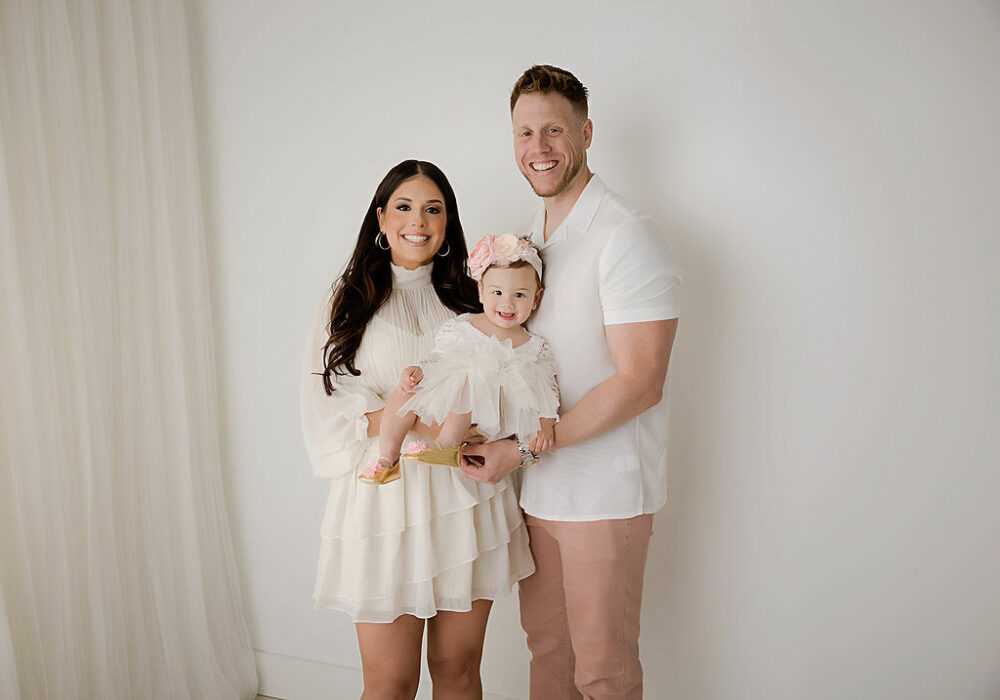 Man and woman posing with their toddler daughter, holding her in both of their arms and smiling for her mermaid first birthday session in Southampton, New Jersey.