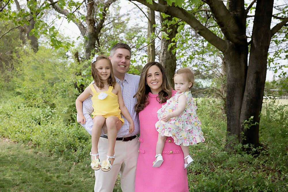 A man and woman standing outdoors and posing with their two daughters for a rainbow pastel first birthday session in Southampton, New Jersey.