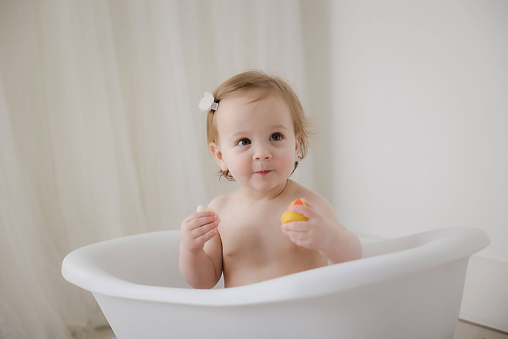 A close-up portrait of a toddler girl playing with a rubber ducky as she sit in bathtub photography prop for her first birthday photo shoot taken in Westampton, New Jersey.