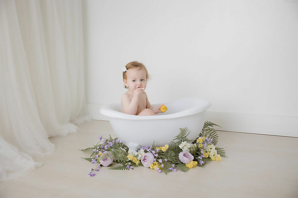 A toddler girl sitting in bathtub photography prop eating a cookie during her birthday cake pic photos taken during her rainbow pastel first birthday session in Medford, New Jersey.