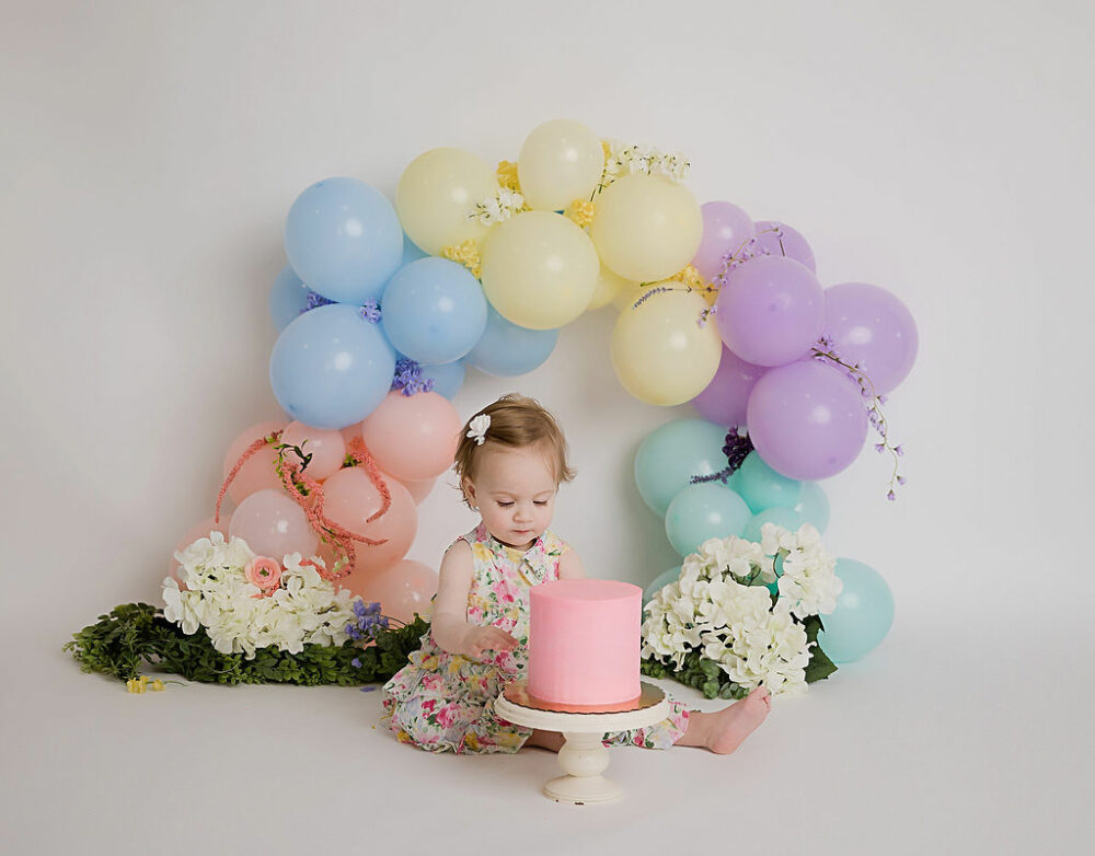 Lifestyle photo of a toddler girl looking at her first birthday cake with a balloon garland backdrop with greenery for her rainbow pastel first birthday session in Camden, New Jersey.