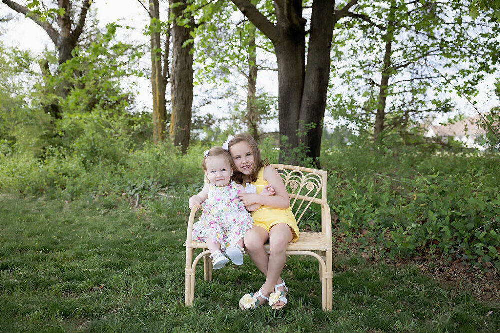 Bling portrait of two sisters sitting outdoors on a small bench smiling for a rainbow pastel first birthday session in Tabernacle, New Jersey.