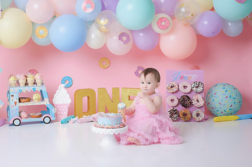A portrait photo of a toddler girl in a cute dress, sitting on the floor and eating a cake against a pink decorated backdrop for her sweet one first birthday session in Westampton, New Jersey.
