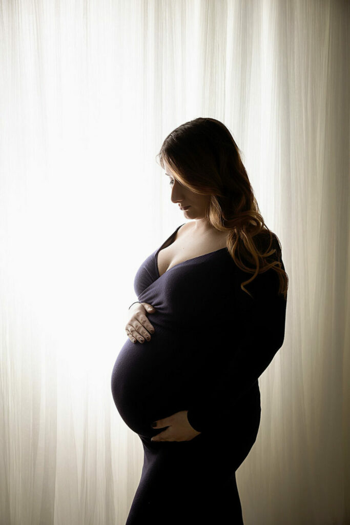 A woman standing and holding her belly as she stands in a 45 degree angle wearing long dress for her classy maternity pics in Westampton, New Jersey.