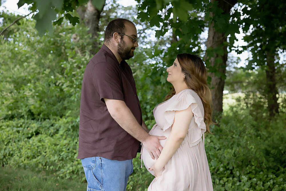 A couple facing each other during an outdoor maternity session looking at each other lovingly while they both hold her belly for a classy pregnancy photo shoot in Cherry Hill, New Jersey.