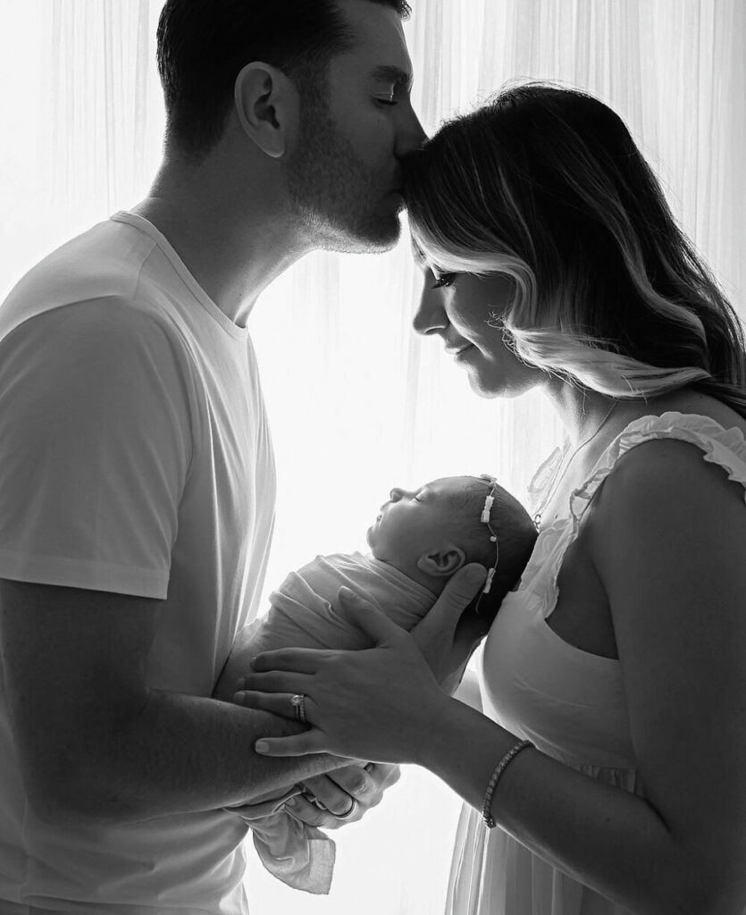 Black and white portrait of a man and a woman facing each other and he is kissing her for head well they both hold their newborn in between them for their newborn portraits taken in Southampton, New Jersey.