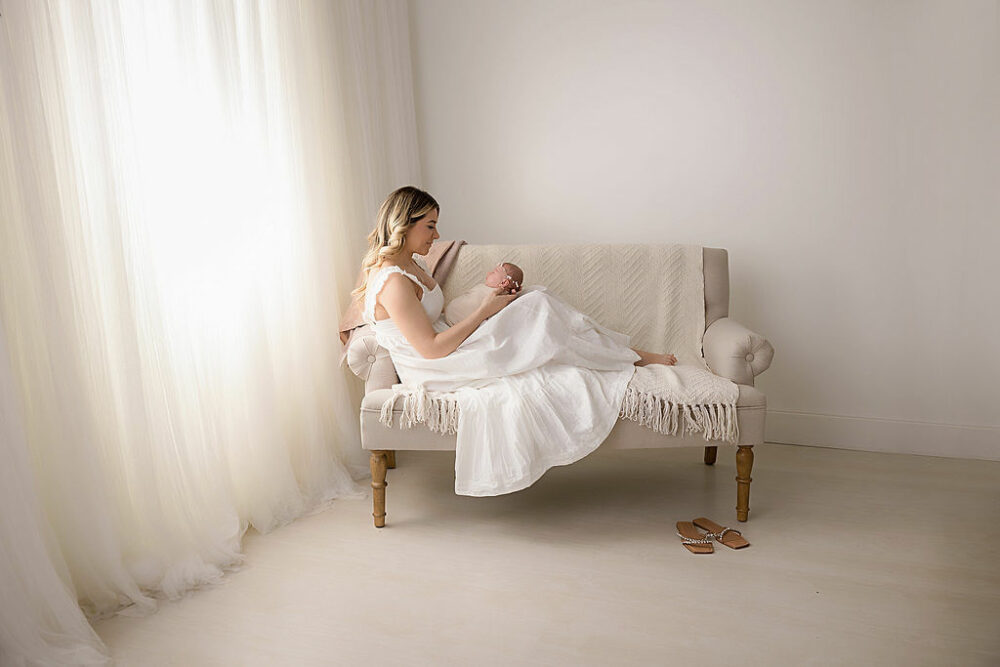 A mommy-and-me portrait of a woman sitting on loveseat with her newborn on her lap looking at her loving me for their In-studio rosy newborn session in Eastampton, New Jersey.
