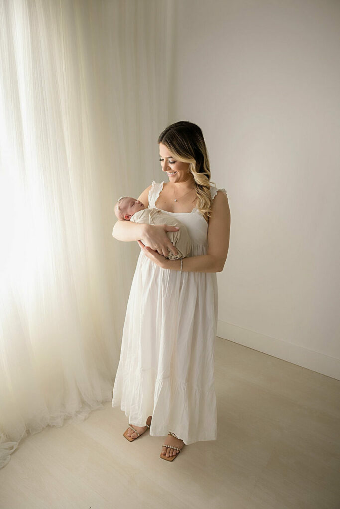 It woman standing and smiling while she holds her baby sleeping in her arms wearing a beautiful dress against a light and bright backdrop for a in-studio rosy newborn session in Deptford, New Jersey.