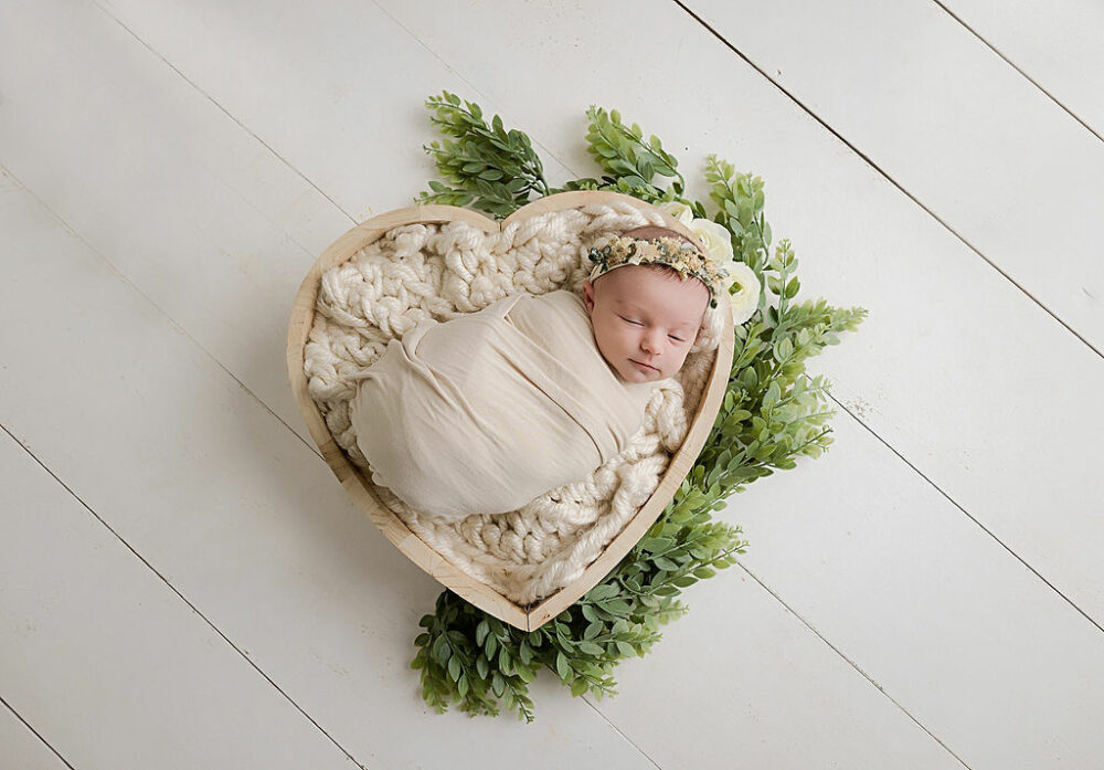 A infant sleeping on textured blanket that is in heart shape photography prop adorned with greenery, swaddled and wearing headband for her baby photo shoot ideas in Wrightstown, New Jersey.