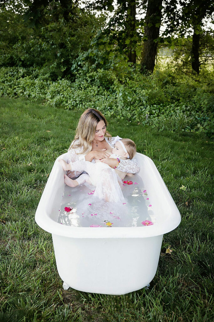 A woman sitting in a bathtub filled with milk and water and flower petals feeding her infant for an outdoor family photo shoot in Deptford, New Jersey.