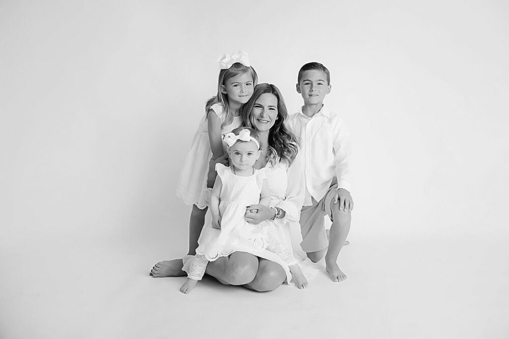 A black-and-white photo of a mother sitting on the floor with her three children standing around her smiling and lightly posed for their lifestyle family photo shoot in Wrightstown, New Jersey.