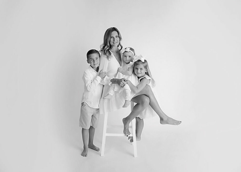 A black-and-white picture of a woman sitting on a stool surrounded by her three children wearing monochromatic colors and lightly posed for a family studio photo shoot in Cherry Hill, New Jersey.